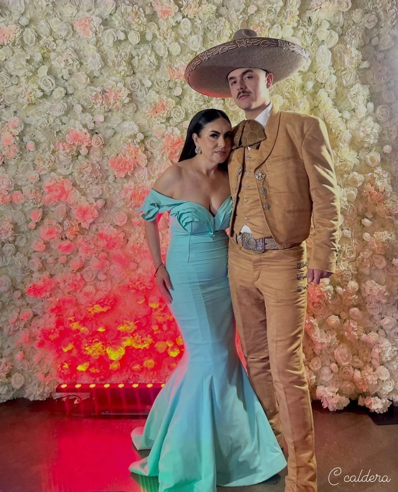 Couple in a teal gown and charro suit posing at the flower wall